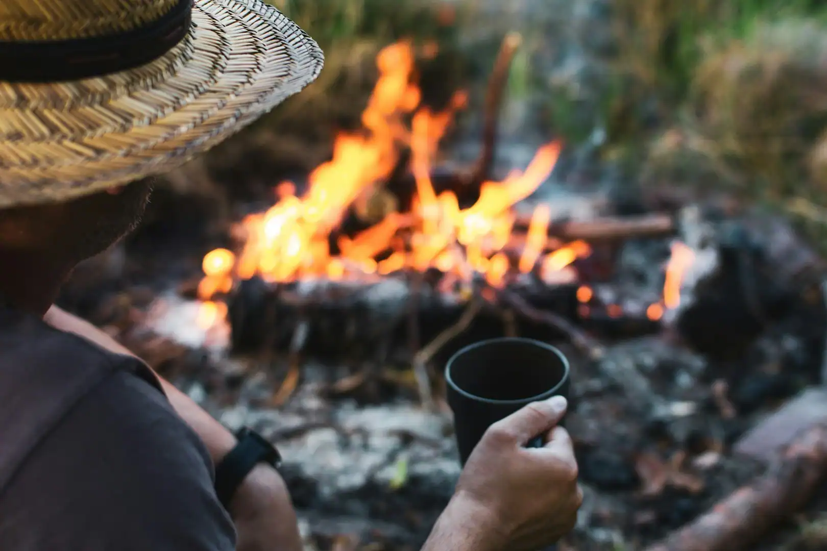 Person sitting by campfire with a cup, wearing a straw hat.